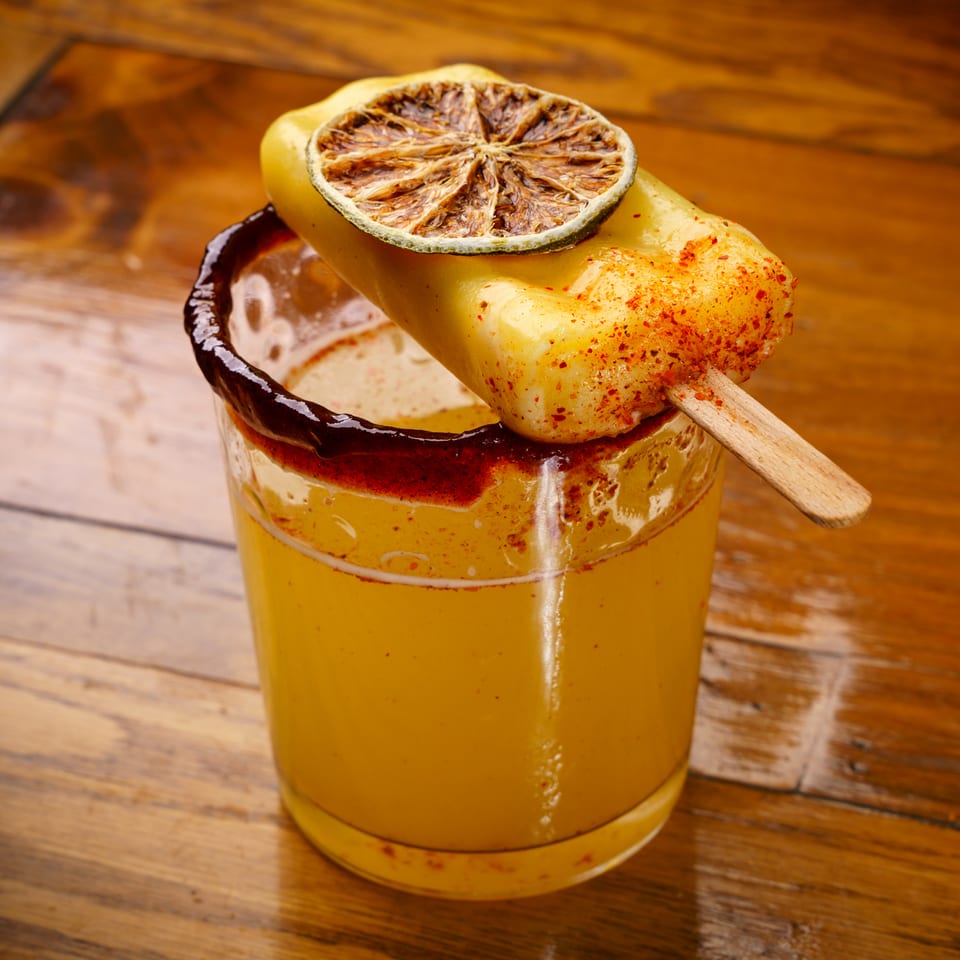Chili-rimmed yellow drink with a spicy popsicle and dried lime garnish on a wooden table.