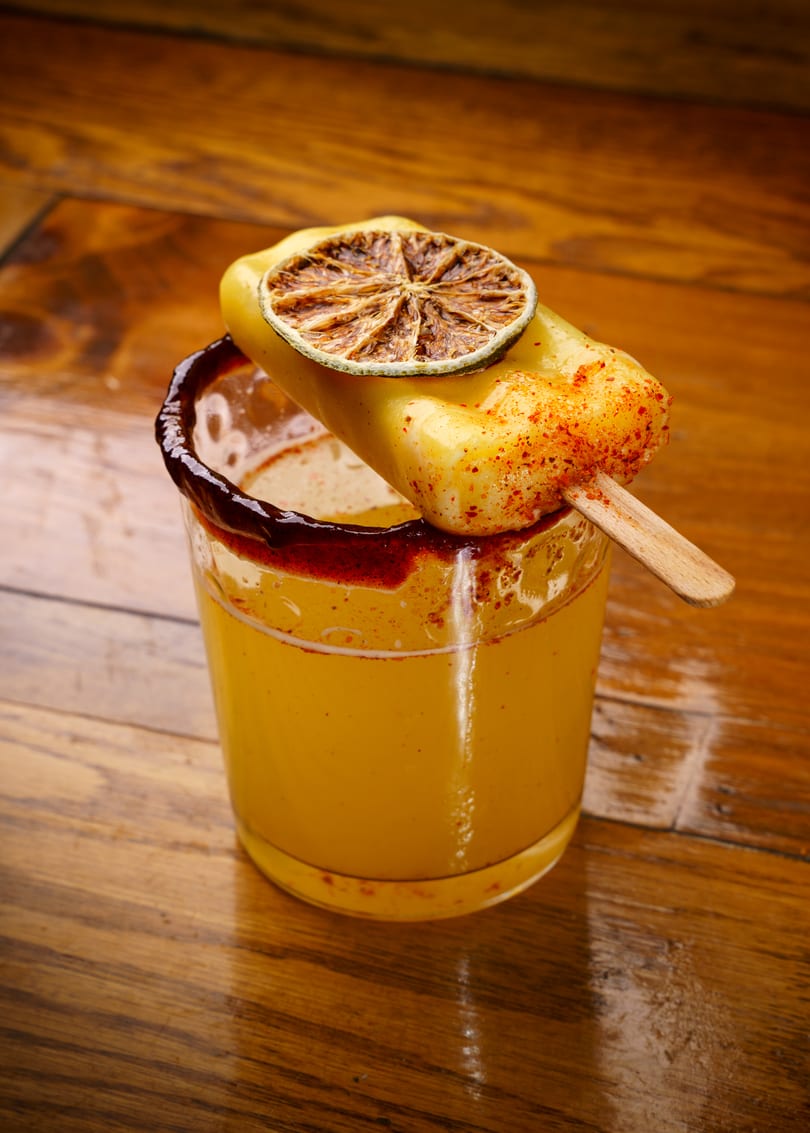 A yellow drink in a chili-rimmed glass, topped with a chili-dusted popsicle and dried lime.
