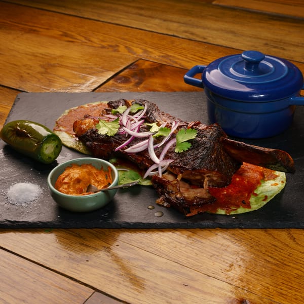 Tender braised meat with red onions, cilantro, jalapeño, sauce, and blue pot on a slate.