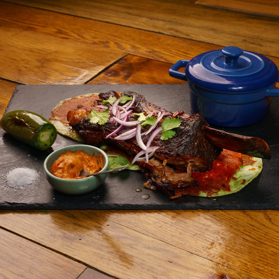 Tender braised meat with red onions, cilantro, jalapeño, sauce, and blue pot on a slate.