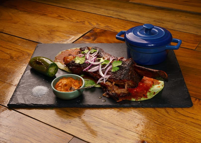 Braised short rib with red onions, cilantro, jalapeno, sauce, and a blue pot on a slate board.