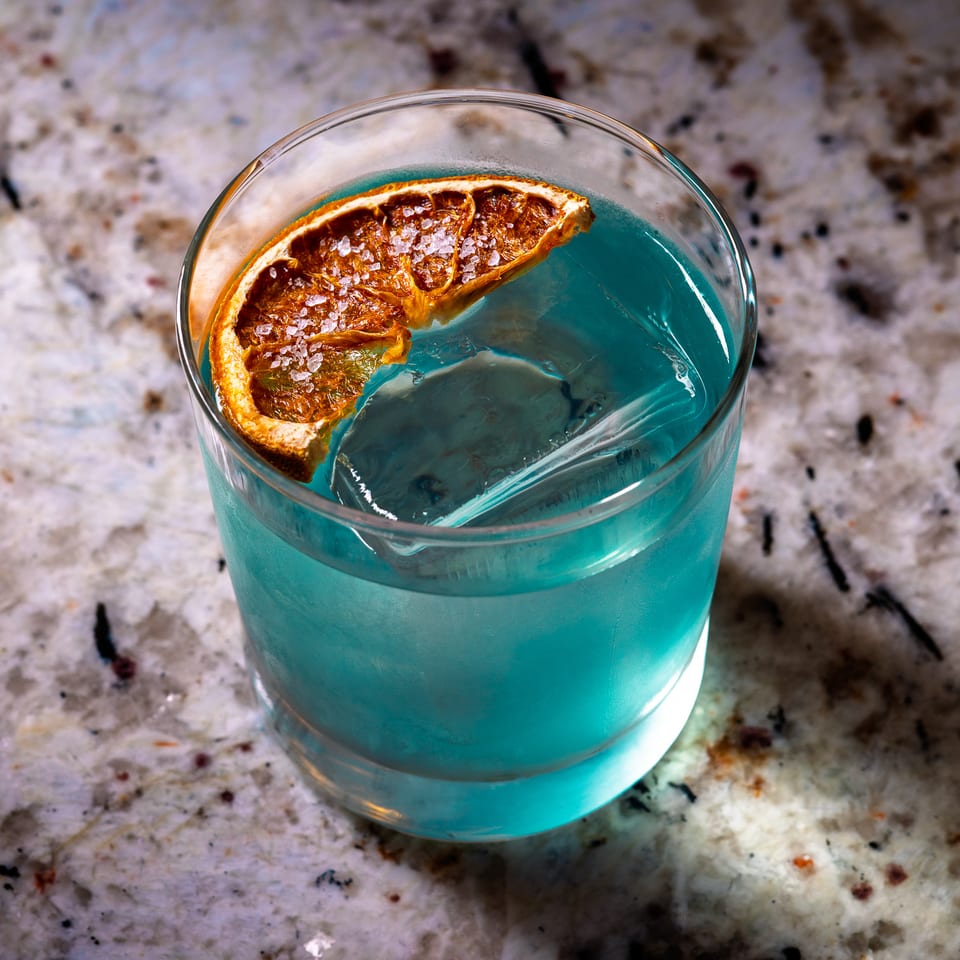 Blue cocktail with a large ice cube and a salted dried orange slice on a speckled counter.