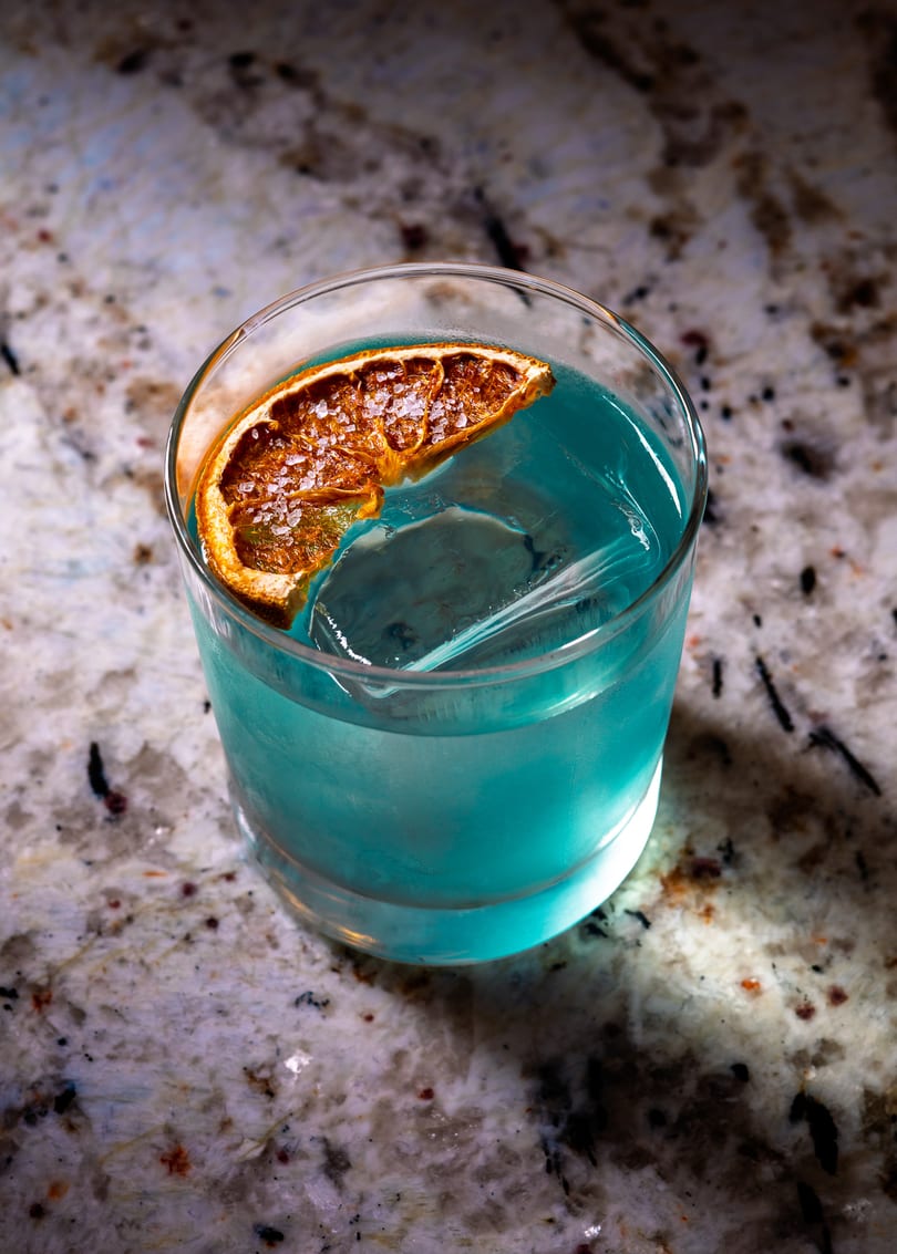 Blue cocktail with a large ice cube and a salted, dried orange slice on a granite surface.