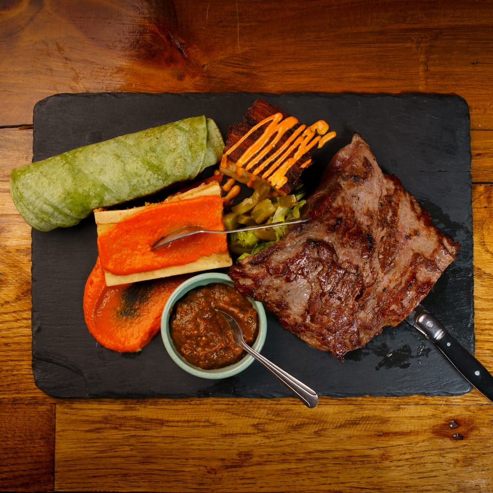 Grilled steak, green wrap, bone marrow, and sauces served on a black slate board.