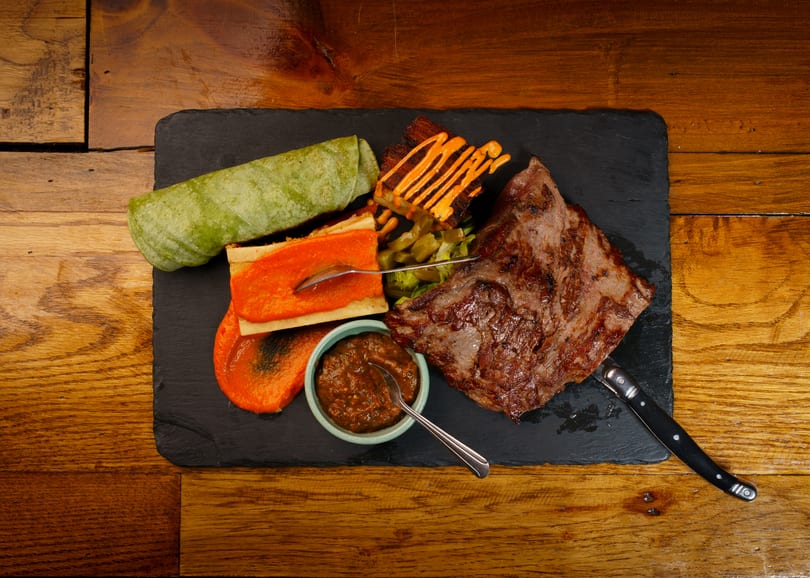 Grilled steak, green wrap, bone marrow, and sauce on a black slate against a wooden table.