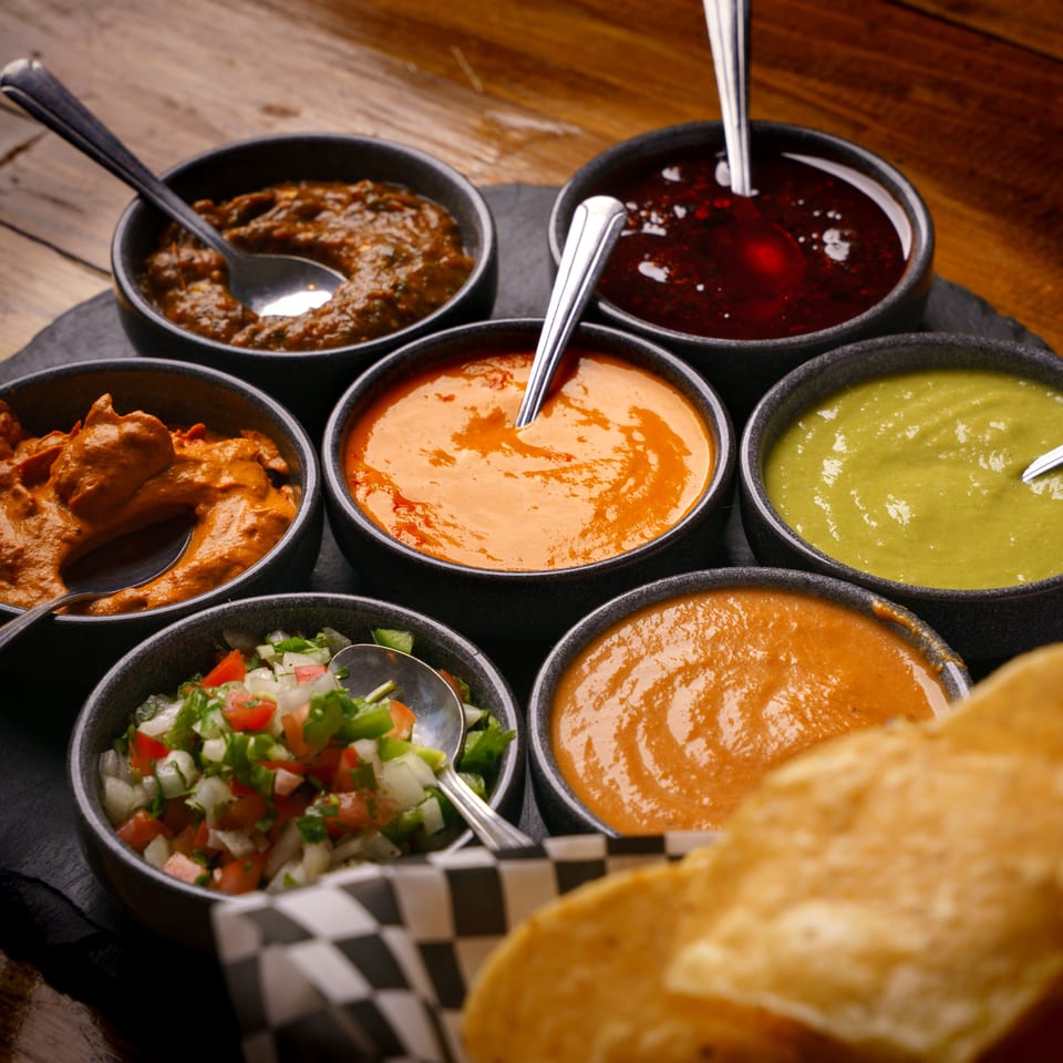 Seven bowls of vibrant salsas and dips with tortilla chips on a dark wooden table.