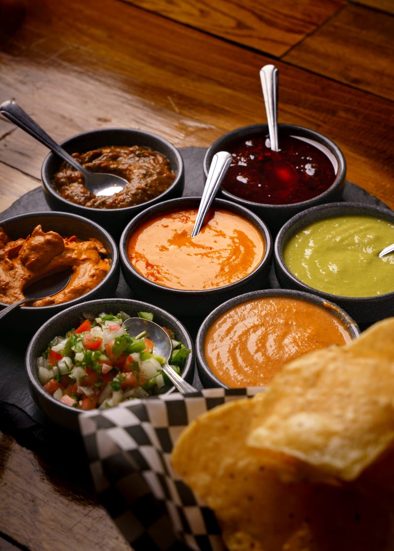Seven bowls of colorful sauces and salsas with spoons, alongside a basket of tortilla chips.