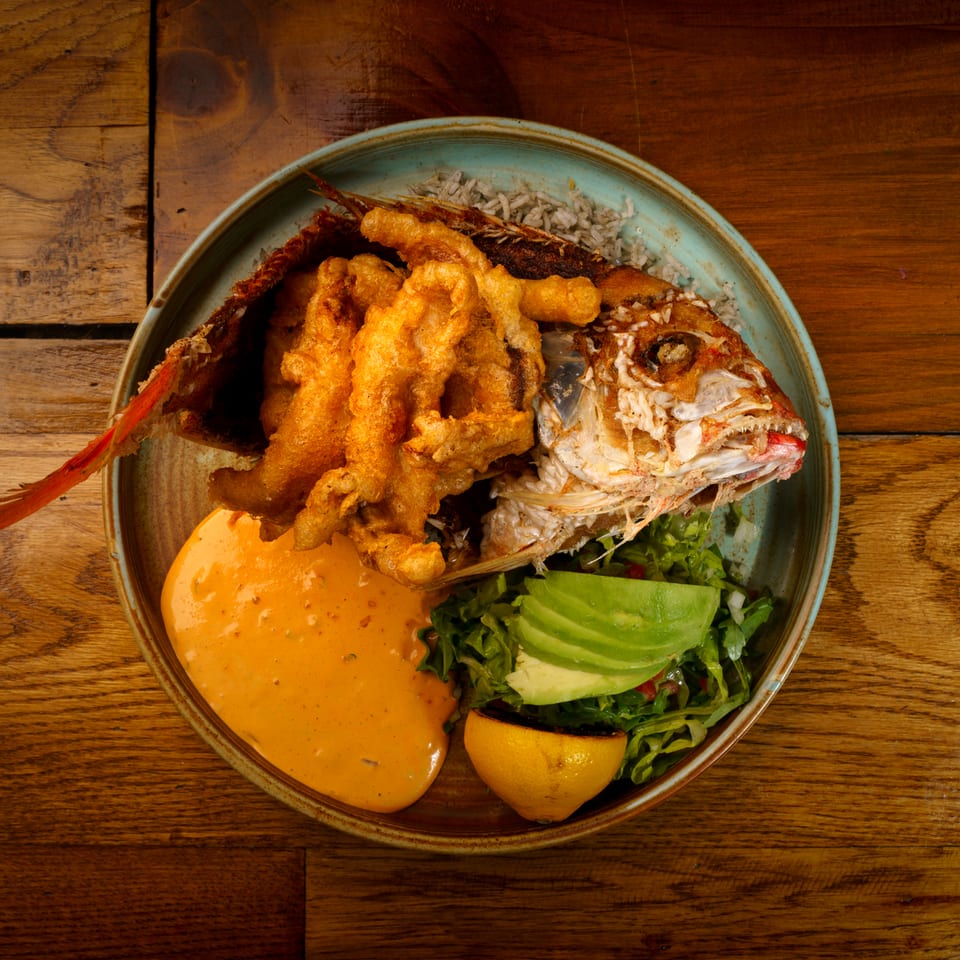 Crispy whole fried fish, orange sauce, green salad with avocado, and grilled lemon on a wooden table.