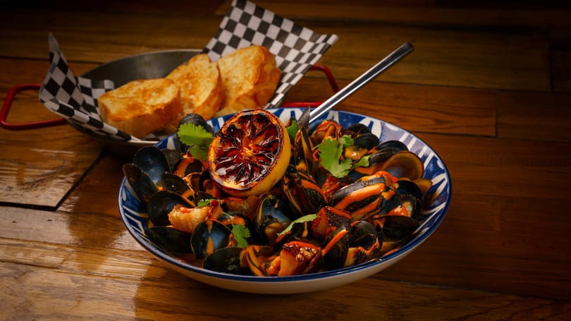 A bowl of mussels in sauce with a grilled lemon half and cilantro, served with toasted bread.