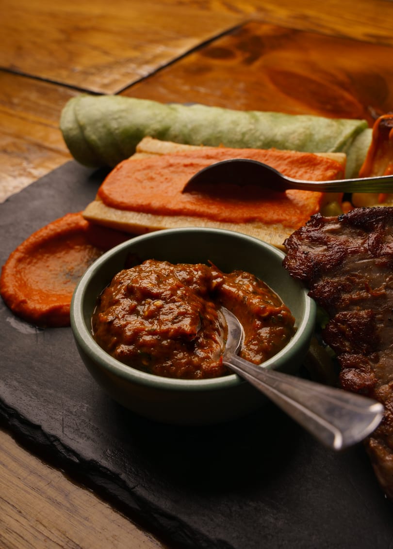 Grilled meat, green wraps, and two bowls of red and brown sauces on a dark slate.